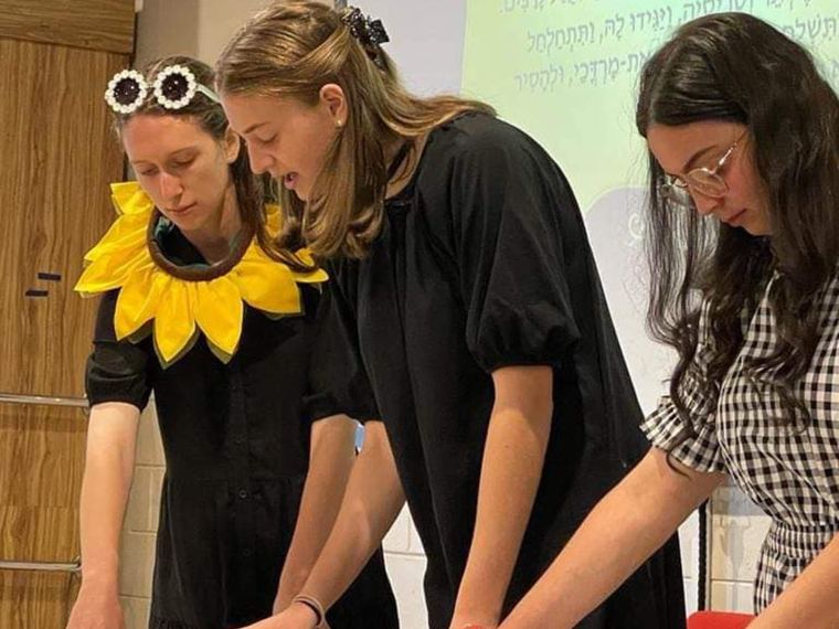 Three women reading from an open scroll on a table, one wearing a Purim costume