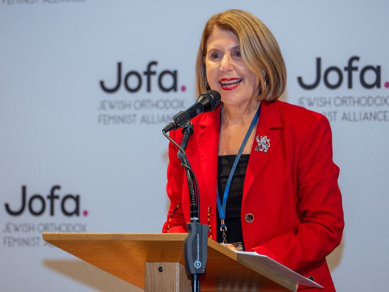 A woman in a red blazer speaking at a podium in front of a Jofa backdrop